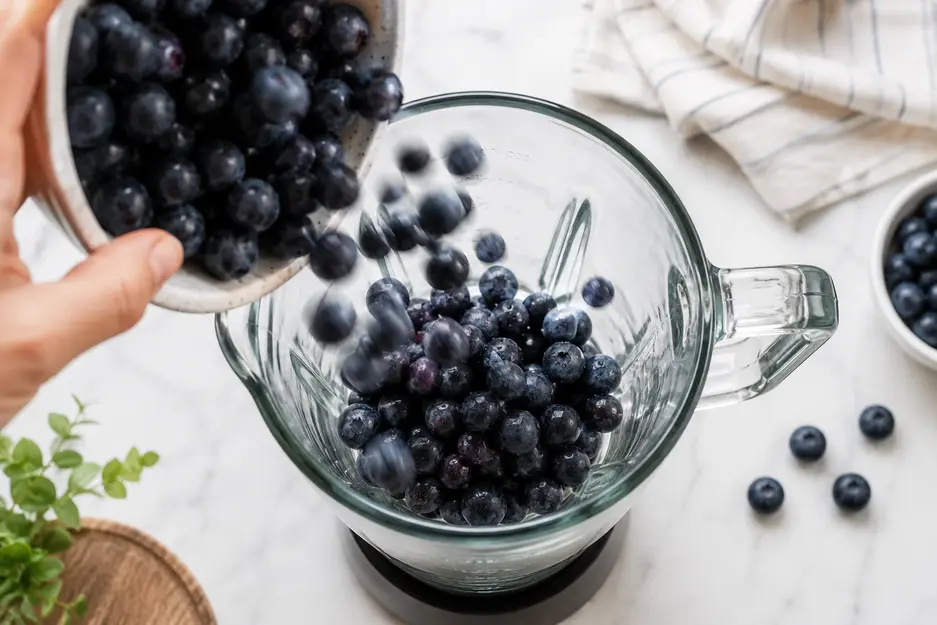 Fresh blueberries being poured into a blender to make homemade blueberry puree for baby on a bright white kitchen countertop.