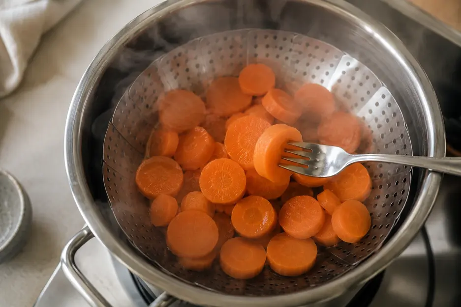 Blender carrot pancakes for baby recipe step showing peeled carrot coins steaming until fork-tender in a stainless-steel steamer basket.