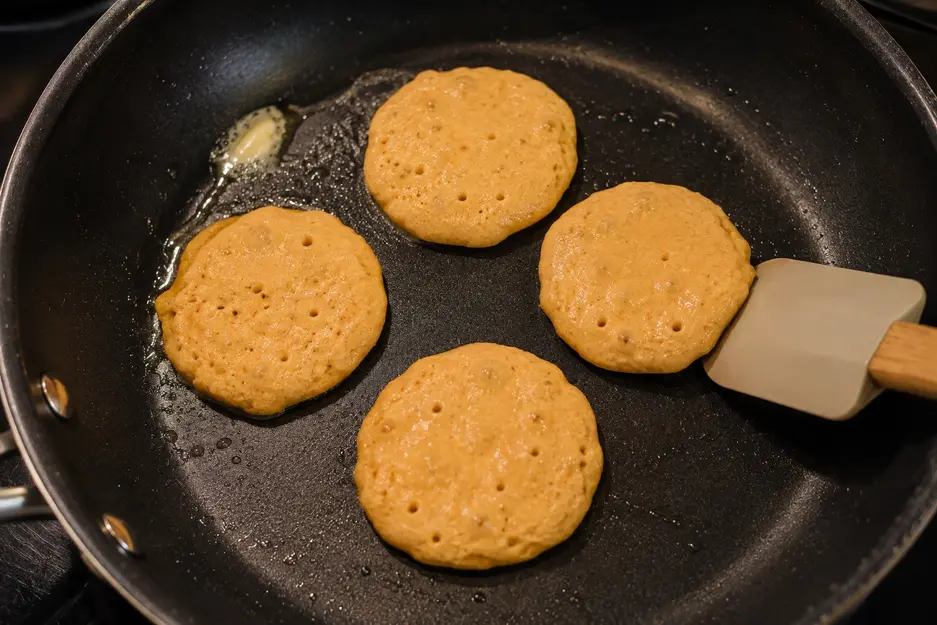 Blender carrot pancakes for baby cooking in a non-stick skillet, with small bubbles and set edges showing the mini pancakes are ready to flip.