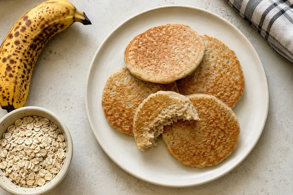 Banana oat pancakes for baby on a cream plate with ripe banana and rolled oats, featuring soft, golden egg-free pancakes with a moist oat texture.