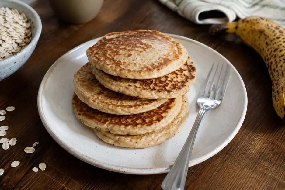Banana oat pancakes for baby stacked on a cream plate with rolled oats and a ripe banana nearby, featuring soft homemade pancakes with natural golden browning.