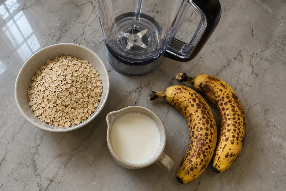 Banana oat pancakes for baby ingredients with rolled oats, two ripe spotted bananas, and milk arranged beside a blender on a kitchen counter.