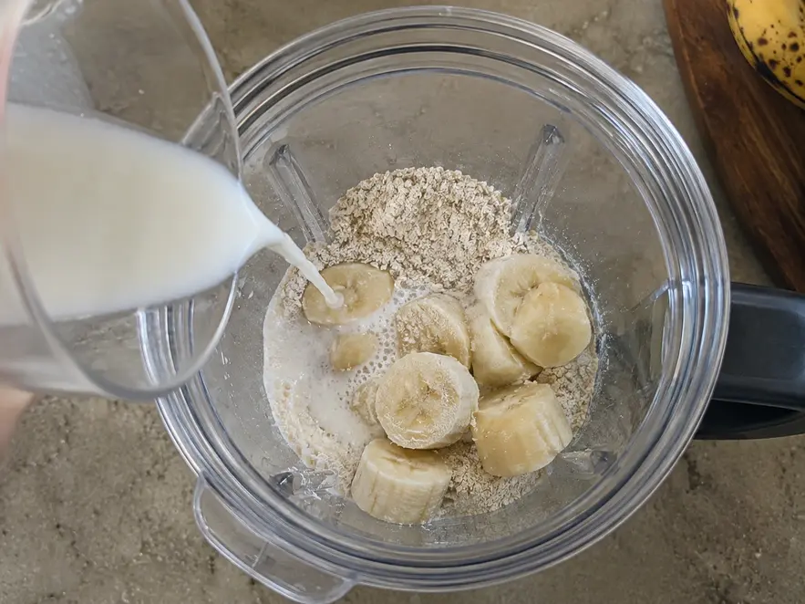 Banana oat pancakes for baby preparation showing banana chunks, oat flour, and milk being poured from a measuring cup into a clear blender.