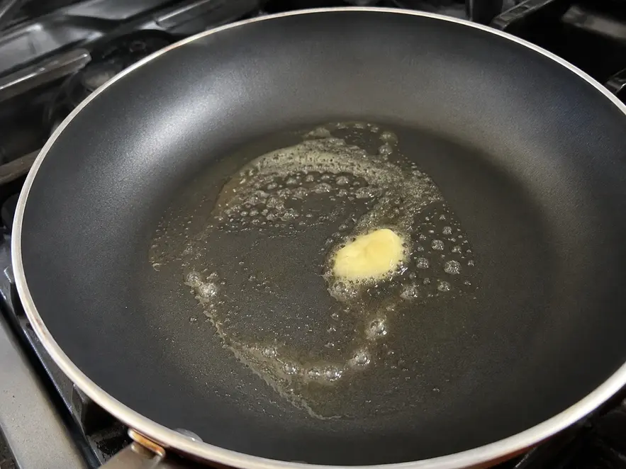 Banana oat pancakes for baby preparation showing a non-stick skillet on the stovetop with a small amount of butter melting across the surface before cooking.