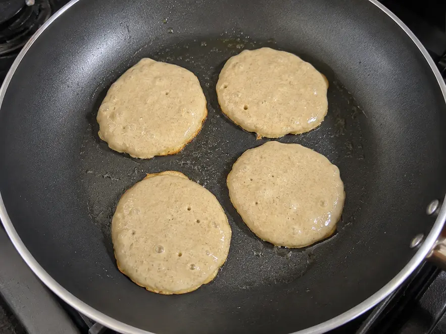 Banana oat pancakes for baby cooking in a non-stick skillet, showing four small homemade pancakes with natural uneven edges and bubbles forming on top.