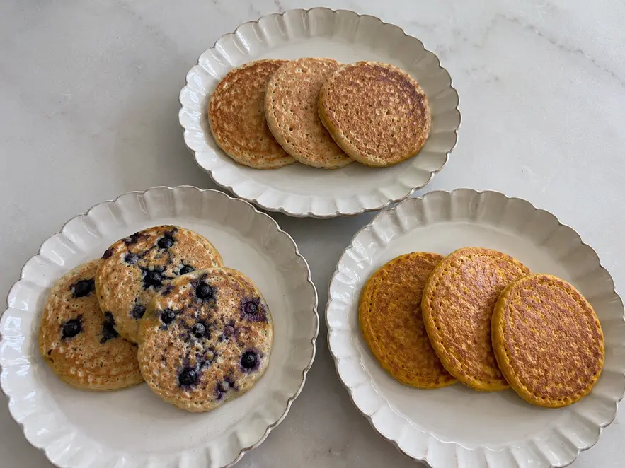 Banana oat pancakes for baby shown in three homemade variations: plain golden pancakes, blueberry pancakes, and pumpkin pancakes served on matching cream ceramic plates.