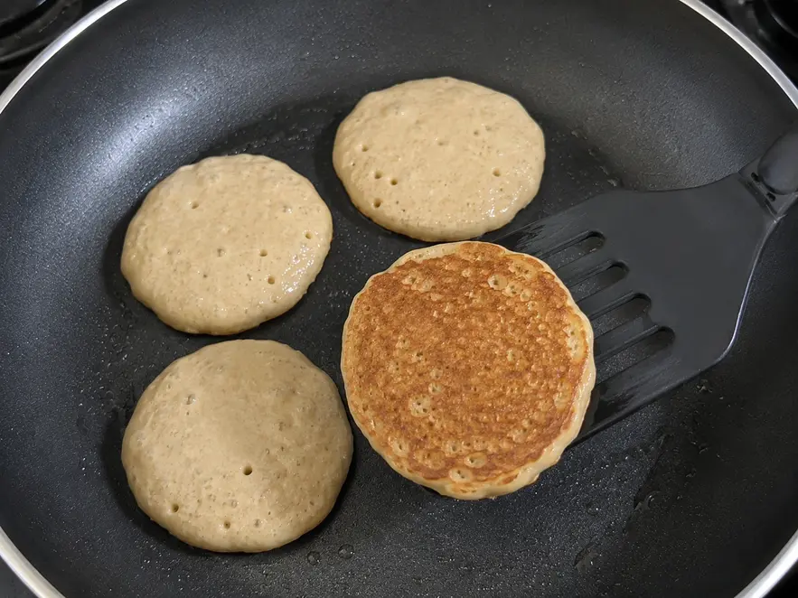 Banana oat pancakes for baby cooking in a non-stick skillet, with one pancake being flipped to reveal a naturally golden-brown underside while three pancakes cook nearby.