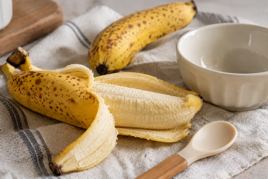 Ripe banana with brown speckles partly peeled beside a cream bowl and baby spoon, ready for homemade banana baby food combinations.