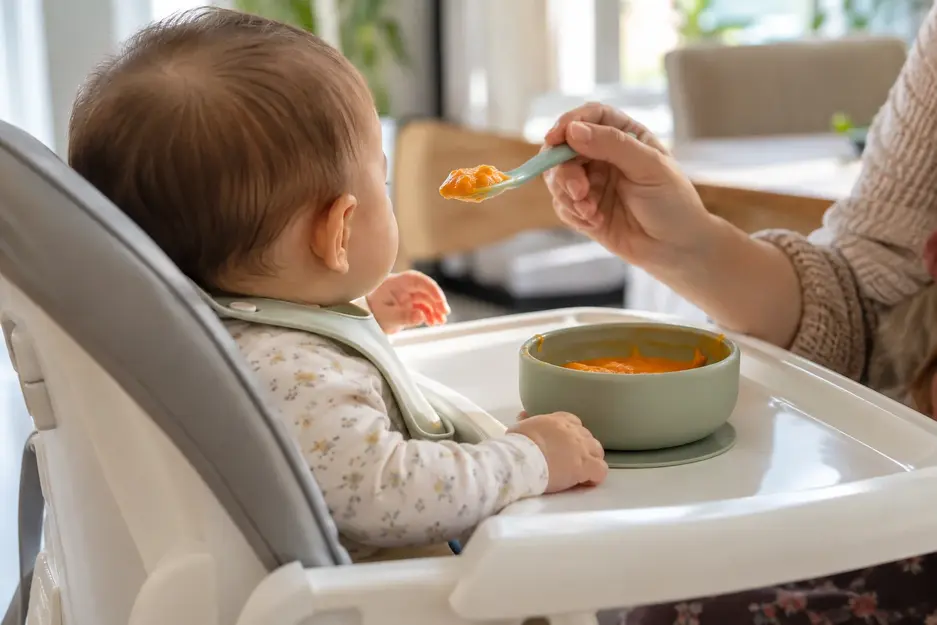 Baby food weekly meal plan for 6–9 months showing a baby seated upright in a high chair while being fed smooth vegetable puree.