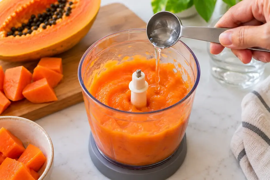 Adding a teaspoon of water to smooth papaya puree in a blender to adjust the texture of homemade papaya puree for baby.