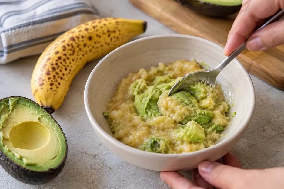 Adult hand gently mixing mashed banana and ripe avocado in a cream bowl, creating a soft, creamy texture for homemade banana baby food combinations.