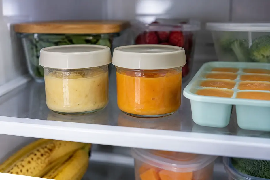 Banana baby food combinations stored in two covered glass containers with pale yellow and orange puree, alongside a silicone portion tray in the refrigerator.