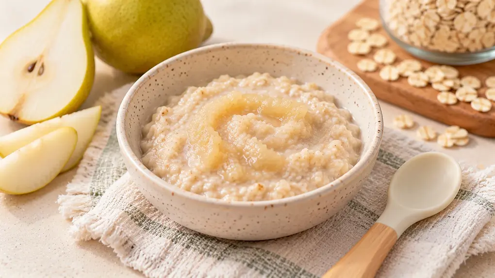 Creamy homemade oatmeal for babies topped with mashed pear puree in a ceramic bowl, served with a baby spoon and fresh pears.