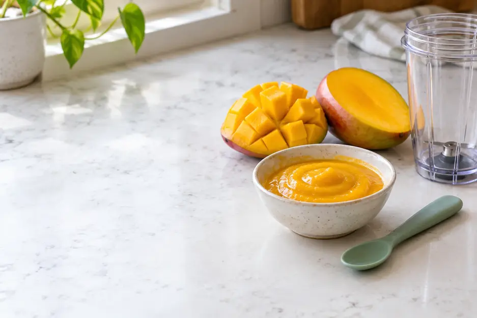 Mango puree for baby in a small ceramic bowl beside a silicone spoon, two ripe mango halves and a clean blender cup on a bright kitchen counter.