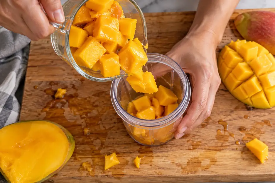 Adult hands tip ripe mango chunks from a measuring cup into an open mini blender cup to prepare mango puree for baby.