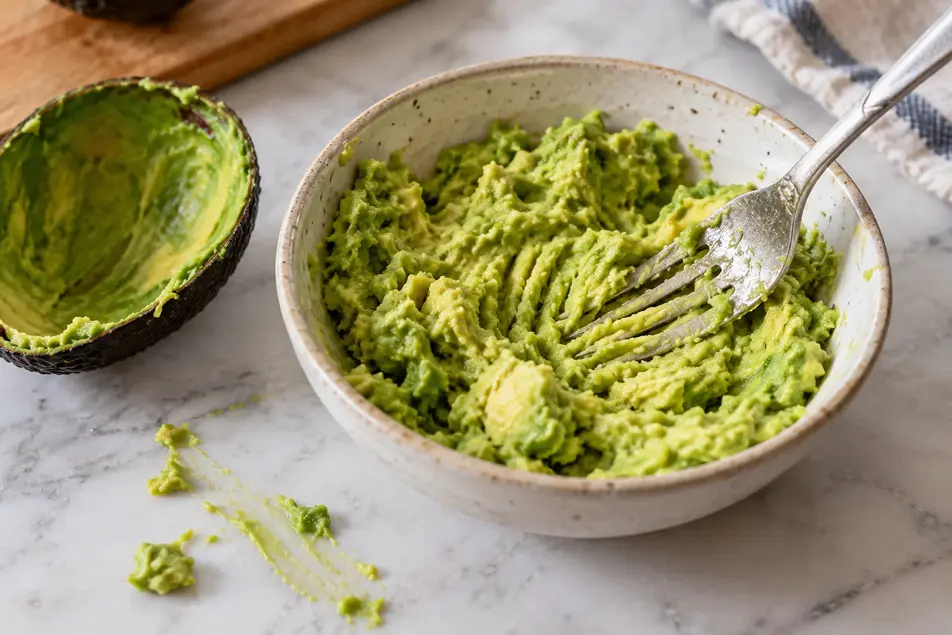 Mashing ripe avocado with a fork in a ceramic bowl to make avocado puree for baby, with an empty avocado shell beside it.